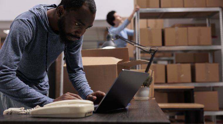 African american employee looking at cargo stock before start preparing customers orders, working in warehouse delivery department. Worker checking shipping detalies on laptop computer