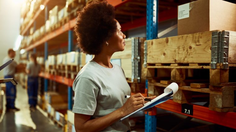 African American worker writing inventory list while checking stock in storage room.