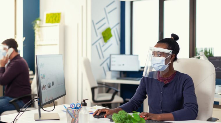 African working at workplace wearing face mask against covid19 as safety precaution. Multiethnic team in new normal business financial office checking reports, analysing datas looking at desktop. New normal.