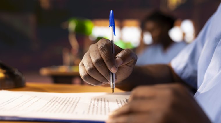 Black male pupil makes notations about practice methods to prepare for an exam, studying at the library to obtain his med school diploma for a medical career. Student does a summary. Close up.