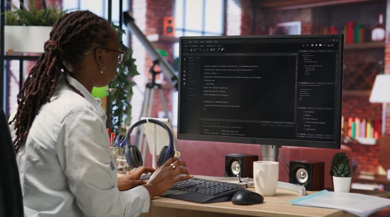 Developer at computer desk coding while listening music, creating software, implementing algorithms. Remote worker with headphones on at home working with code editor, building applications, camera B