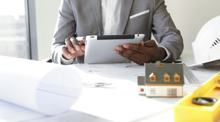 Selective focus. Design and architecture. Cropped shot of African architect holding digital tablet, developing new real estate project, sitting at desk with architectural tools and scale model house