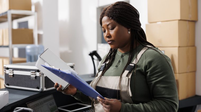 Small business worker analyzing merchandise logistics, checking customers orders on computer, preparing packages for delivery. Stockroom employee wearing industrial overall in warehouse