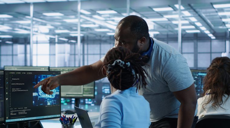 Manager coming to technician desk to give instructions on doing maintenance on artificial intelligence neural networks. Supervisor overseeing worker using AI system software, camera A