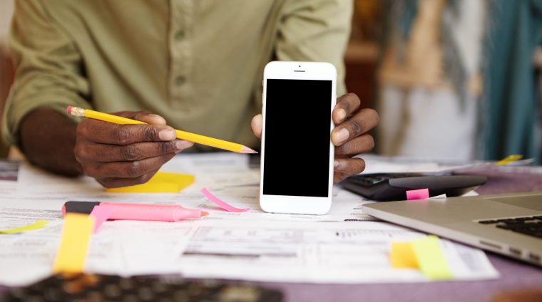 Unrecognizable African man holding yellow pencil, pointing it at blank screen smart phone in his hand while calculating family expenses and paying utility bills online, using electronic gadgets